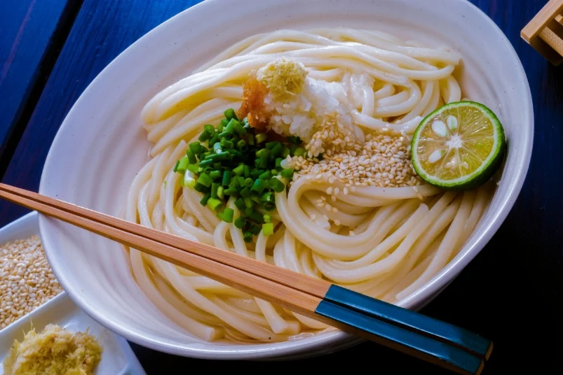 A bowl of freshly prepared Sanuki udon noodles garnished with green onions, sesame seeds, and citrus.