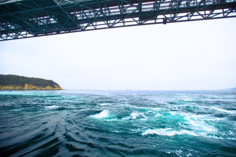 The dynamic Naruto whirlpools churning in the sea beneath a bridge structure.