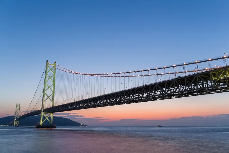 A large suspension bridge illuminated at dusk over a calm sea.