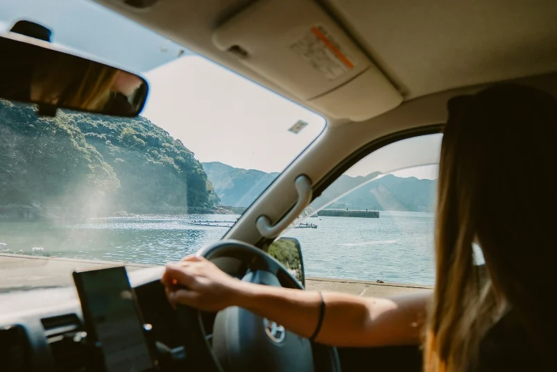 A driver's perspective from inside a Travellers Autobarn campervan, looking out at a bright coastal road and ocean view during a road trip in Japan.
