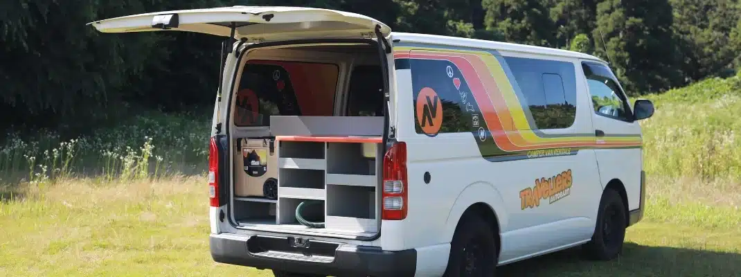 Open rear hatch of a Travellers Autobarn campervan showing built-in storage shelves and a portable cooler box.