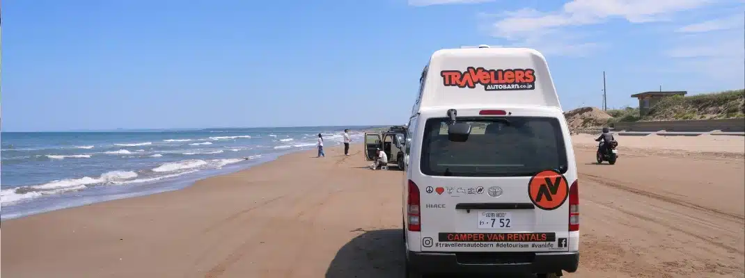 A white Travellers Autobarn rental campervan parked on a sandy beach next to the ocean waves in Japan under a clear blue sky.