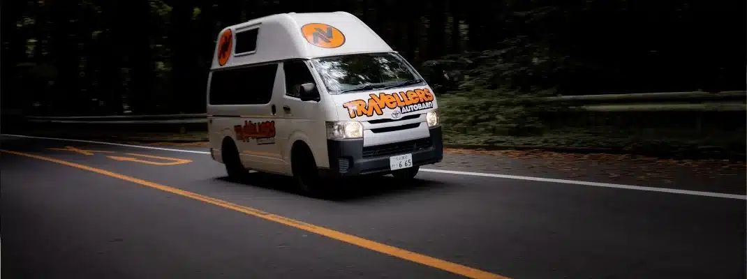 A white Travellers Autobarn campervan driving along a road surrounded by green trees