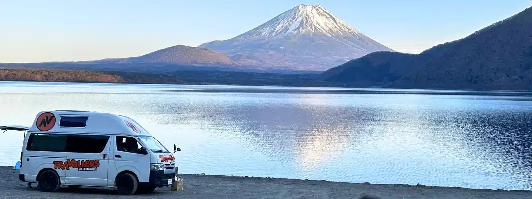A white campervan parked by Lake Motosu with snow-capped Mount Fuji reflecting in the water