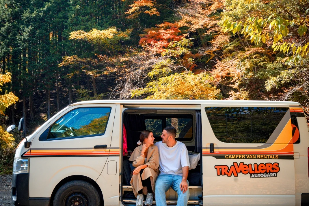 A happy young couple sitting inside the open side door of a white Travellers Autobarn rental campervan, smiling at each other with a backdrop of colorful autumn mountain trees.