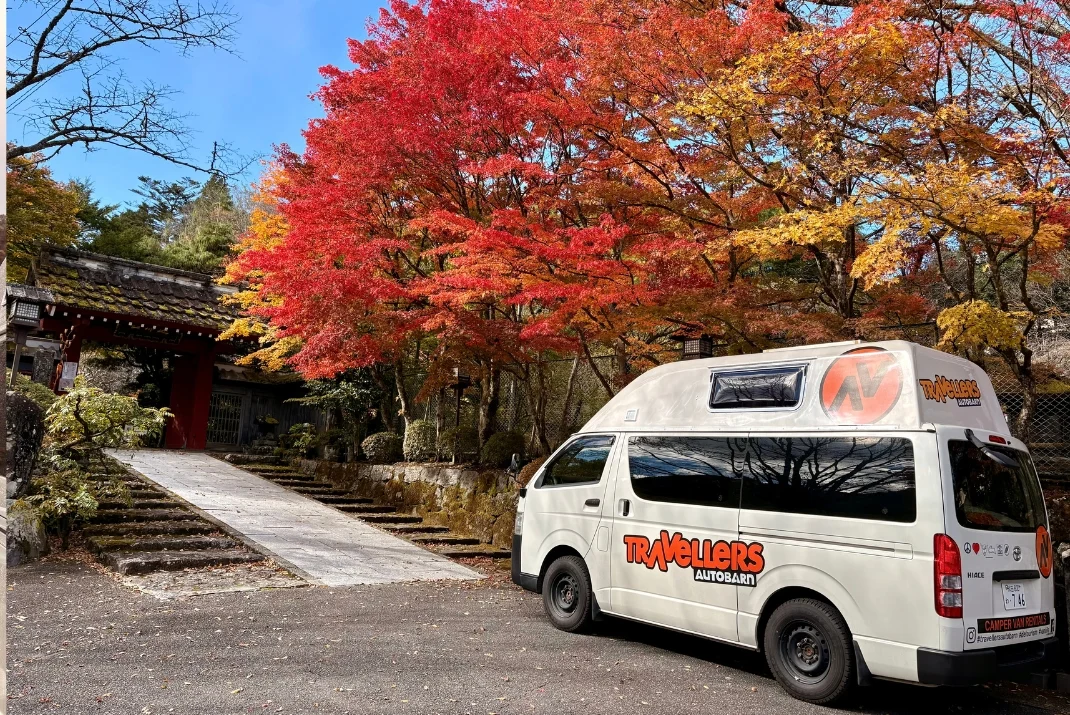 A Travellers Autobarn rental campervan parked near a traditional historic gate and brilliant red autumn trees in Nikko during a autumn road trip.