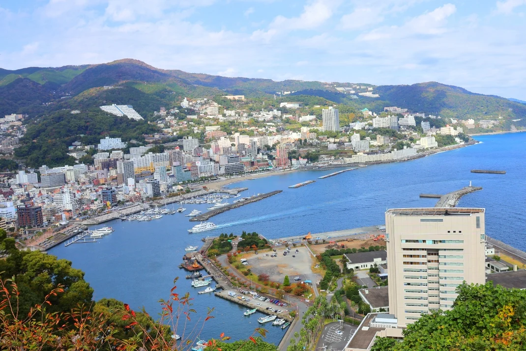 High-angle panoramic view of Atami's coastal city skyline, harbor filled with boats, and blue ocean bay surrounded by mountains.