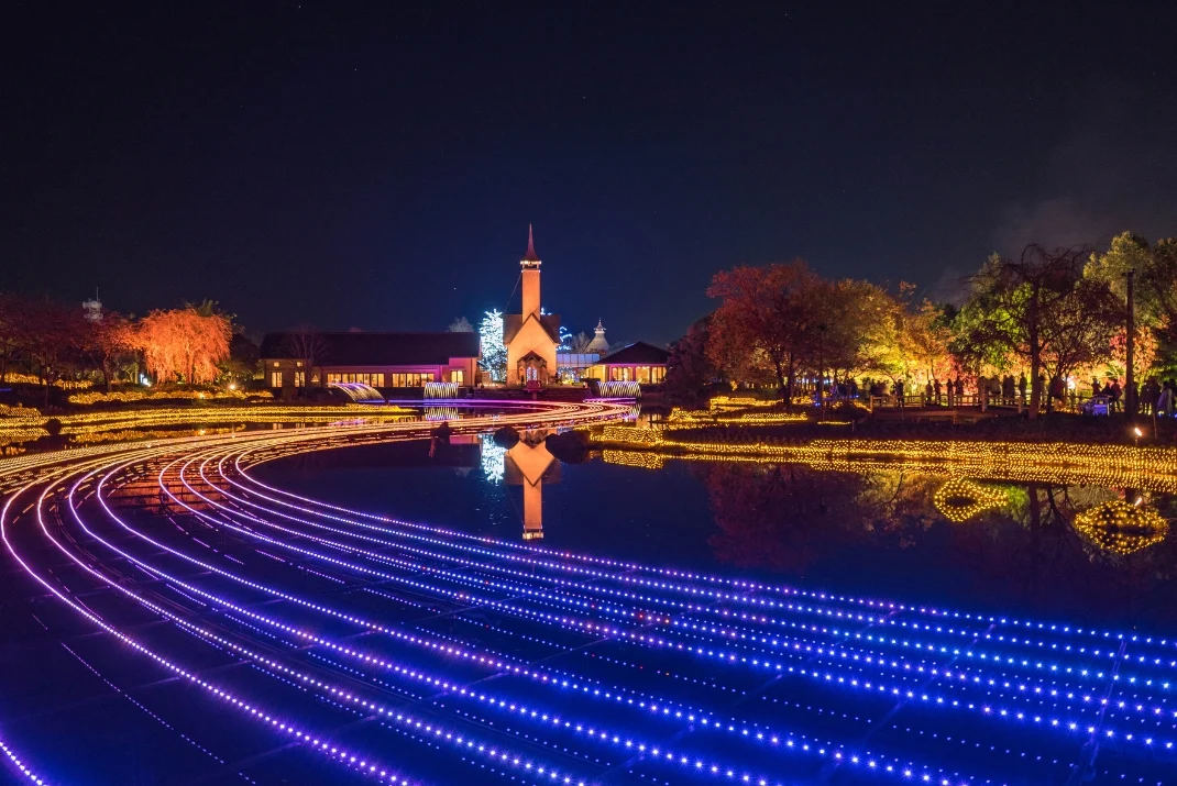 Vibrant purple and blue LED light installations curving across a pond at night at Nabana no Sato, with a chapel reflection in the water.