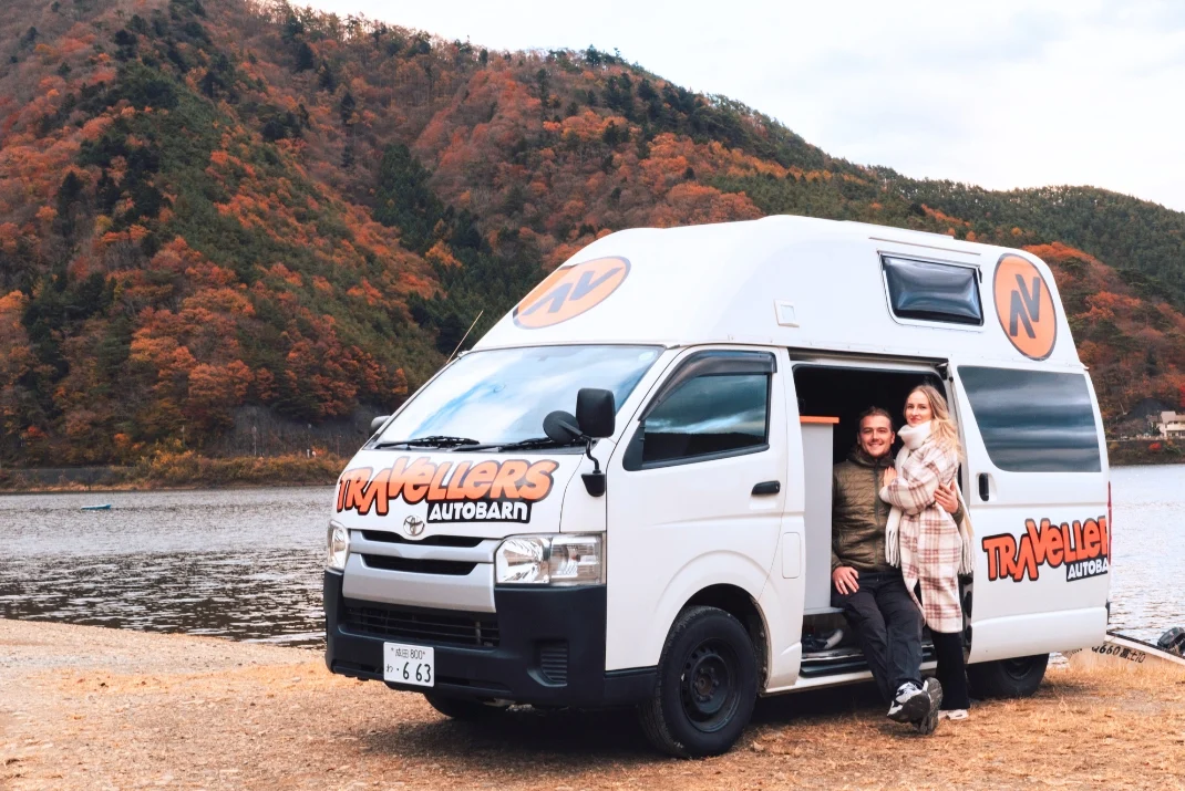 A smiling couple sitting at the side door of a white Travellers Autobarn Japan campervan parked by a lake, with autumn-colored foliage covering the mountainside in the background.