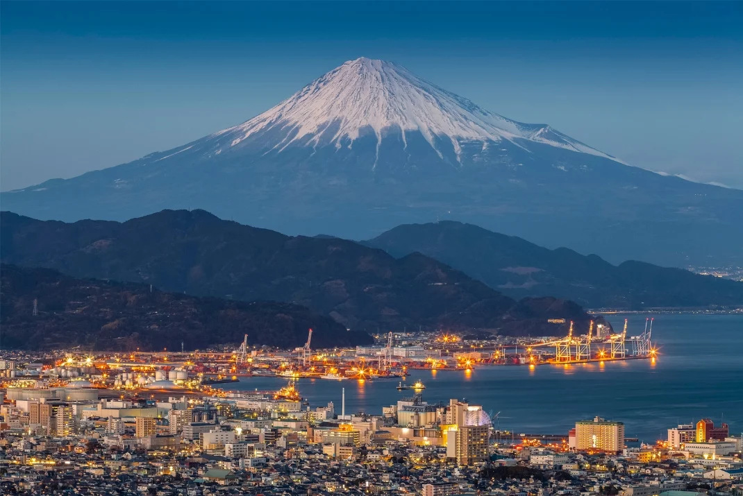 High-angle night view of the illuminated Shimizu Port city skyline with snow-capped Mount Fuji rising majestically in the background.