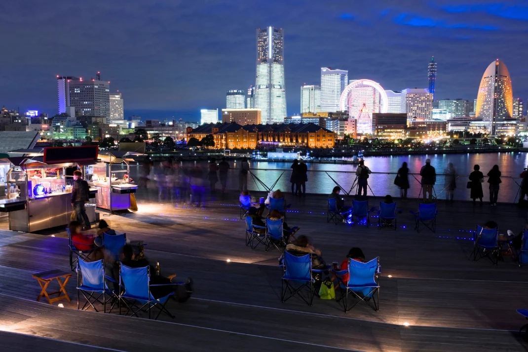 Night view of Yokohama Minato Mirai skyline with illuminated skyscrapers, Ferris wheel, and people relaxing on an open deck area in the foreground.